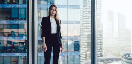 Half length portrait of pretty blonde business woman in trendy elegant formal wear standing near window in office interior, confident beautiful 30s female executive manager in suit looking at camera