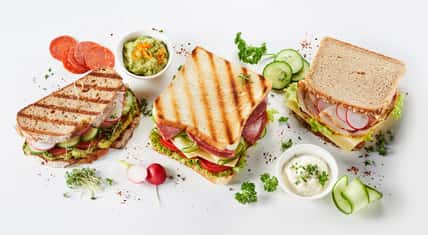 Trio of gourmet sandwiches on assorted bread with meat, cheese, guacamole and pesto fillings surrounded by ingredients and seasoning on white in a panorama banner