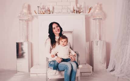 Smiling mother sitting with baby girl in armchair at home. Looking at camera. Motherhood.