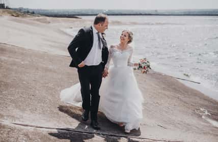 Cheerful elderly groom with bride walkiing along beach holding hands.