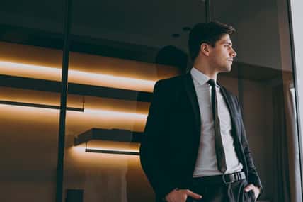 Young serious successful employee agent business man corporate lawyer 20s wear formal black suit shirt tie look aside illuminated indoors apartment dark wall. Achievement career lifestyle concept.