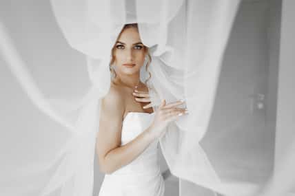 A woman in a white wedding dress is standing in front of a curtain. She is wearing a veil and has her arms crossed. The image has a romantic and intimate mood