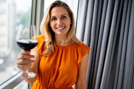 Beautiful young woman with glass of red wine standing near window