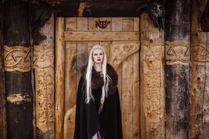 Outdoors portrait of beautiful furious scandinavian warrior ginger woman in a traditional clothes with fur collar, with sword in her hand and wooden Viking Village view on the background.