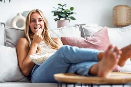 Shot of laughing young woman watching a movie on tv while eating popcorn lying on the couch at home