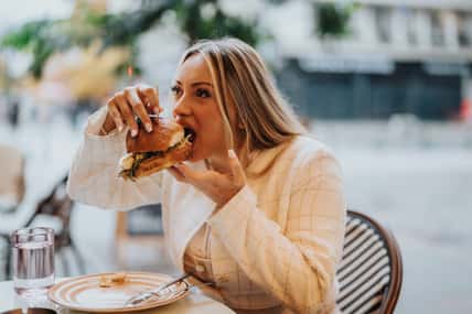 A woman is savoring a large sandwich while dining outdoors at a cozy cafe. The setting conveys a relaxed and enjoyable atmosphere, perfect for casual dining and appreciating delicious food.