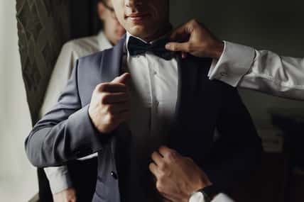 stylish groomsmen helping happy groom getting ready in the morning for wedding ceremony. luxury man in suit in room. space for text. wedding day.