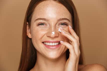 Beauty image of pretty shirtless woman smiling and applying face cream isolated over beige background