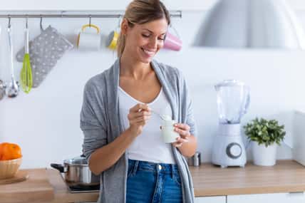 Shot of pretty young woman eating yogurt while standing in the kitchen at home.