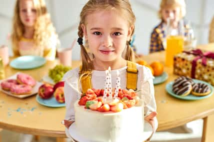 girl with birthday cake in hands, she looks at camera, celebrating with friends, kids behind table in the background