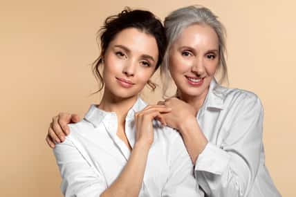 Charming beautiful mother and daughter in white casual shirt studio face forward portrait. Affectionate senior woman hugging adorable young girl shoulder from back. Adult child touching mom hand