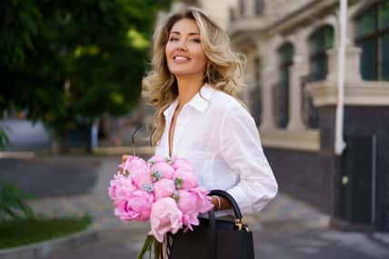 Portrait of elegant beautiful cheerful blond woman in white blouse and silk skirt , holding pink boquet with pions flowers. Stylish hand bag and sunglasses.