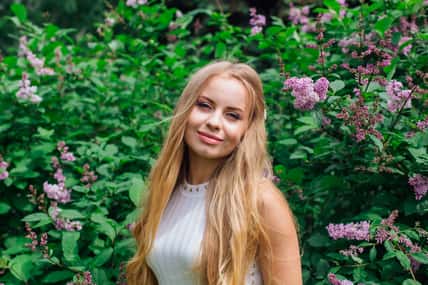 Spring portrait of a charming blond woman wearing beautiful white dress standing next to blooming purple lilac bush.