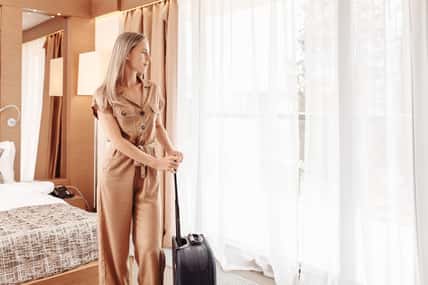 Successful joyful young beautiful well-groomed businesswoman in brown overalls with suitcase settled in luxury room and enjoys magnificent view from window. Young traveler enjoys relaxing in spa hotel