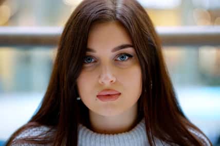 Young woman with long hair and blue eyes looking at camera, blurred background. Handheld closeup portrait of beautiful female model. Concept of beauty