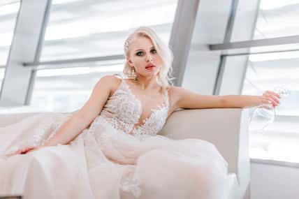 An attractive pensive blonde woman with a creative hairstyle for short hair is sitting on a white sofa in the lobby of the hotel. Art photography. White long wedding dress of the bride.