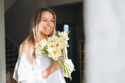 Young beautiful smiling woman forty year with blonde long hair in white shirt with bouquet of yellow flowers in hands near window in bright interior at home