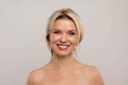Portrait of cheerful attractive half-naked middle aged blonde woman smiling at camera, posing on grey studio background, demonstrating glowing perfect skin, copy space. Cosmetology concept. High quality photo