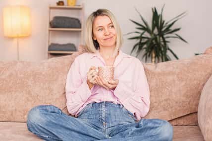 Satisfied middle aged woman is sitting on couch at home with cup of coffee. Smiling adult female of 40 years old is sitting on sofa in living room