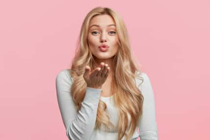 Pretty blonde woman blows passionate kiss into camera, expresses her love, isolated over pink background. Adorable young female model makes air kiss. People, body language and beauty concept