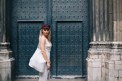 Half length portrait of female traveller with stylish eco bag posing near architecture with ancient doors in historic center, trendy dressed hipster girl in claret cap visiting touristic town