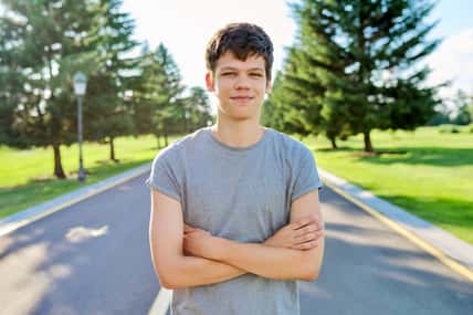 Portrait of a guy 16, 17 years old looking at the camera. Smiling teenager with crossed arms, on the road in the park, summer sunset background
