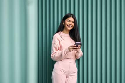 Happy Asian teen girl wearing pink outfit using mobile phone apps outdoors looking at camera. Positive smiling pretty female model holding smartphone laughing standing at blue wall background.