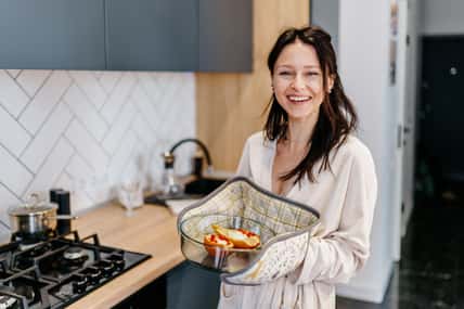Mother in her 40s smiling and holding baked pears in glass dish, standing in modern home kitchen. Joyful moment after home cooking. Concept of healthy lifestyle, positive motherhood, simple pleasures.
