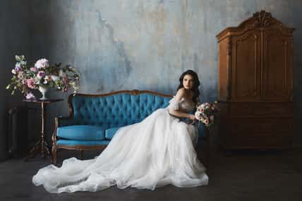 beautiful brunette model girl in stylish and fashionable lace wedding dress with naked shoulders sits on the antique sofa and posing in luxury vintage interior.