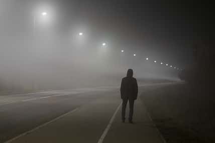 Young man alone slowly walking under white street lights in night. Dark time. Peaceful atmosphere in mist. Foggy air. Back view.