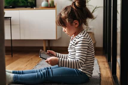 Portrait of little girl in jeans and t-shirt sitting on carpet and holding tablet