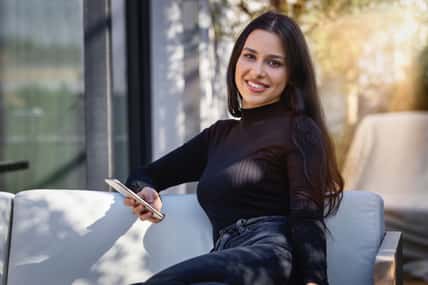Portrait of a young woman with long, black hair sitting on a terrace and holding her mobile phone