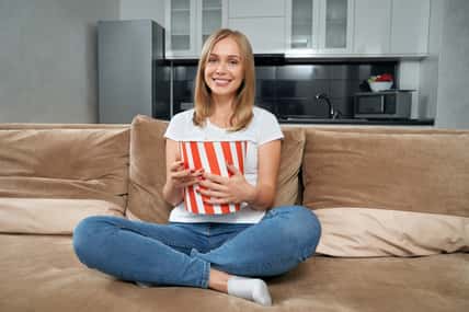 Happy woman in casual outfit sitting with crossed legs on couch with big striped bucket of popcorn. Smiling blonde relaxing at home and watching movie.