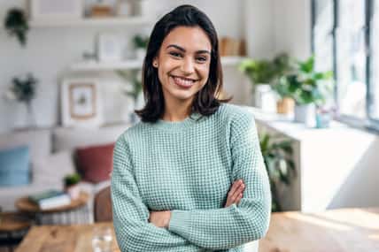 Portrait of beautiful young woman looking at camera while standing in the living room at home.