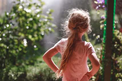 Back view of seven years old blonde girl wearing pyjamas standing outdoor in home garden in the early morning.