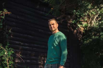 Handsome bearded man in a green sweater posing by log wall and green trees on the sunset. Young eco friendly hipster.