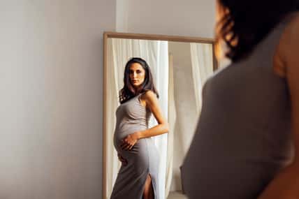 Close up of female portrait of beautiful pregnant in grey dress touching her belly. Reflection of young woman expecting baby in large rectangular mirror. Light color curtains on background.