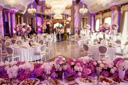 Look from behind dinner table decorated with garland of pink and white flowers at large dinner hall