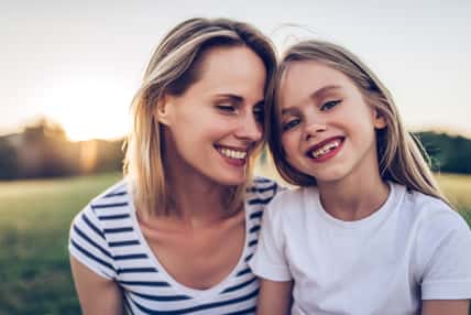 Attractive young woman with her little cute daughter are spending time together outdoors. Mom with daughter in park on a green grass during the sunset.