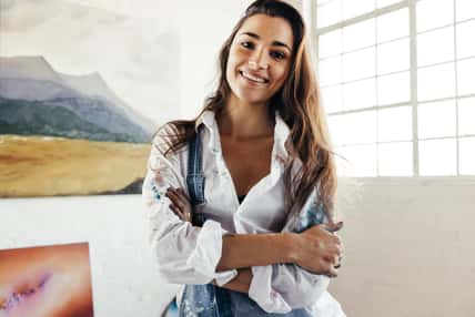Cheerful artist smiling at the camera while standing in front of her paintings. Happy female painter standing with her arms crossed. Creative young woman displaying a collection of her artwork.