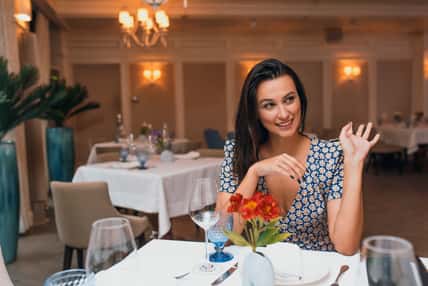 Happy brunette young elegant woman wearing dress sitting in the restaurant waving her hand to her friend. Caucasian female posing in cafeteria and waiting her family for a dinner. Copy space for text