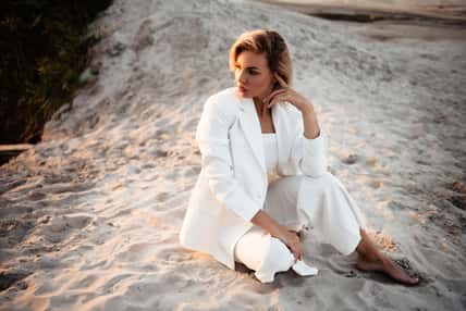 Stunning young woman with blond hair sitting on sand and looking aside during professional photo session on nature. Female model in white suit posing on camera outdoor.