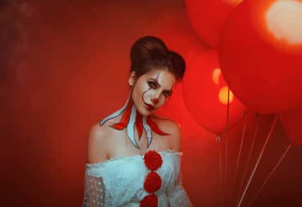 portrait make up young woman crazy smiling face clown in white costume in dark gothic room holding red balloons evil eyes. art creative bright halloween holiday make-up body art painting. smoke party