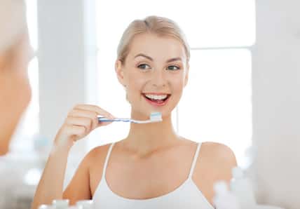 smiling young woman with toothbrush cleaning teeth and looking to mirror at home bathroom