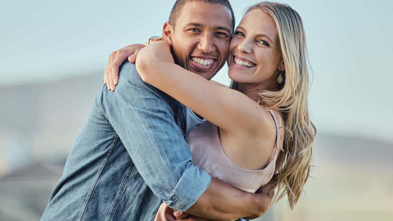 Interracial, happy couple and smile portrait hug of people in summer with happiness or love. Couple, outdoor and smiling for marriage, care or vacation gratitude in sunshine together feeling calm.
