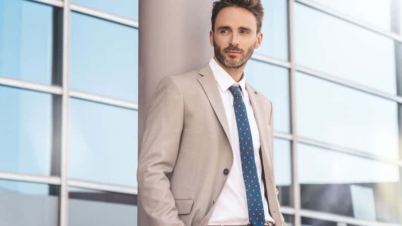 Young, handsome, elegant businessman is standing by the glassy office building wearing a light suit