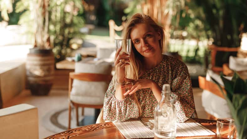 Young happy woman waiting for order at restaurant, look at the camera, hold glass of water in her hands. Beautiful young lady drinking water in restaurant