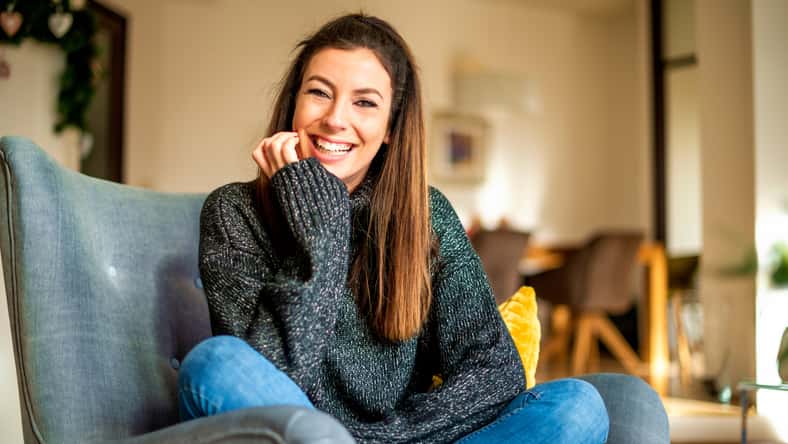 Shot of an attractive smiling woman wearing turtleneck sweater and jeans while relaxing in the armchair at home.