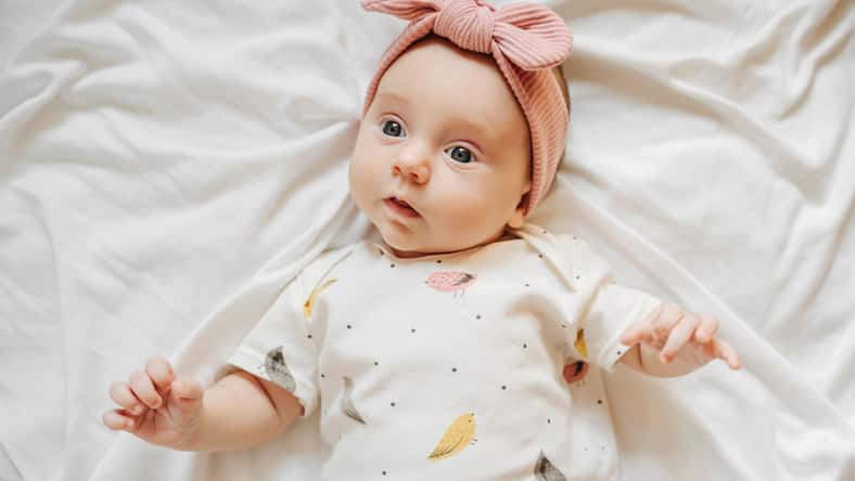 Cute stylish baby in trendy clothes and pale pink headband lying on bed. Fashion baby girl