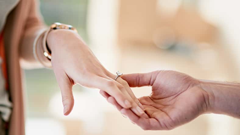 Couple, hands and engagement with ring in home, love and relationship with diamond jewellery on fiancee. Affection, wedding proposal and commitment with partner, romance and apartment together.